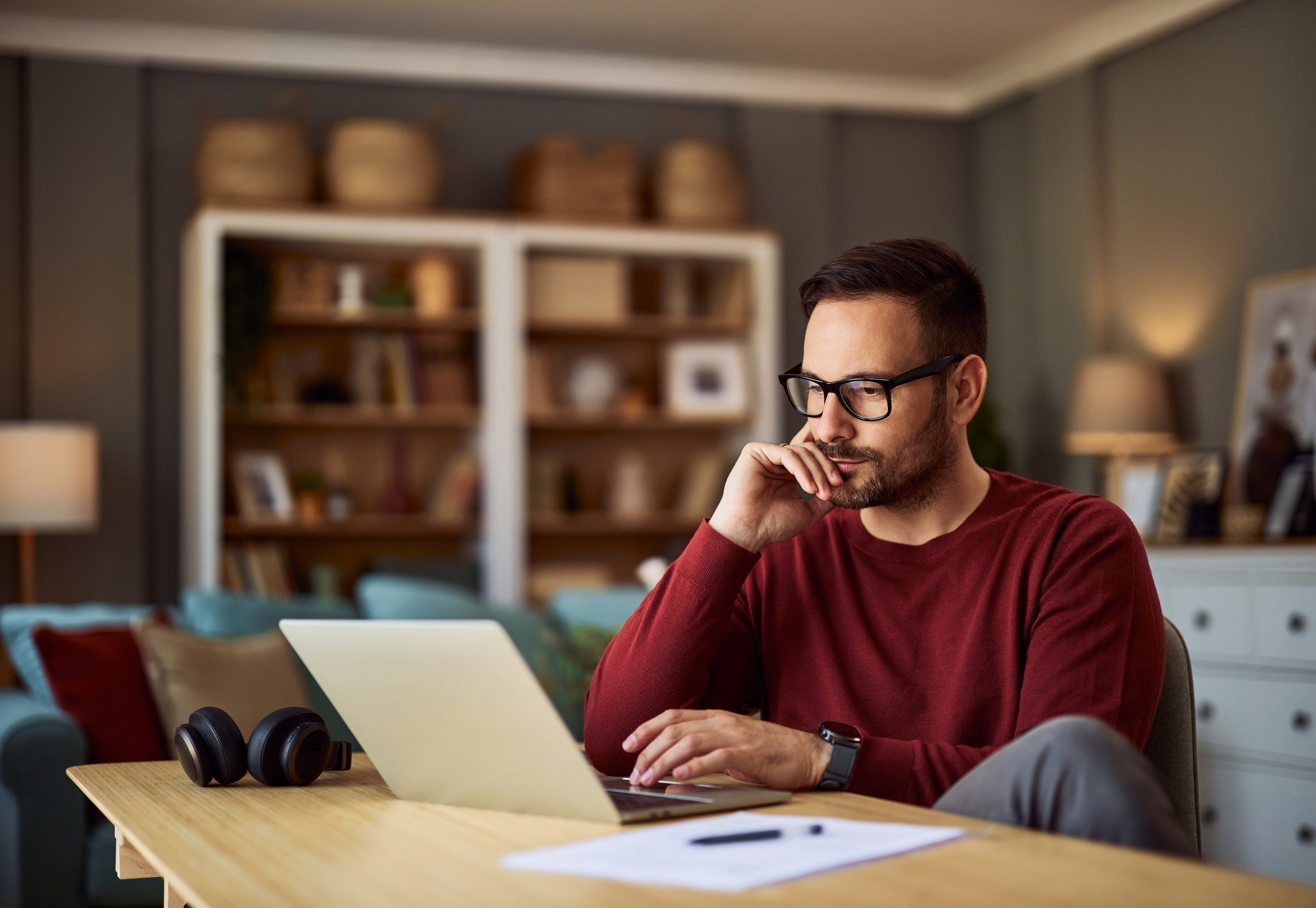 A busy, focused young adult male freelancer sitting at a desk and resting his head on his hand while working on a project on his laptop.